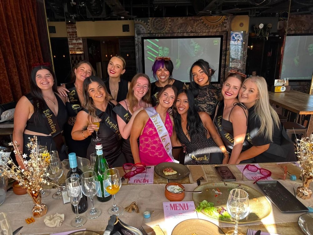 Britt as maid of honour, front left with a glass of bubbles, and the hens group in black and gold sashes around a long lunch table