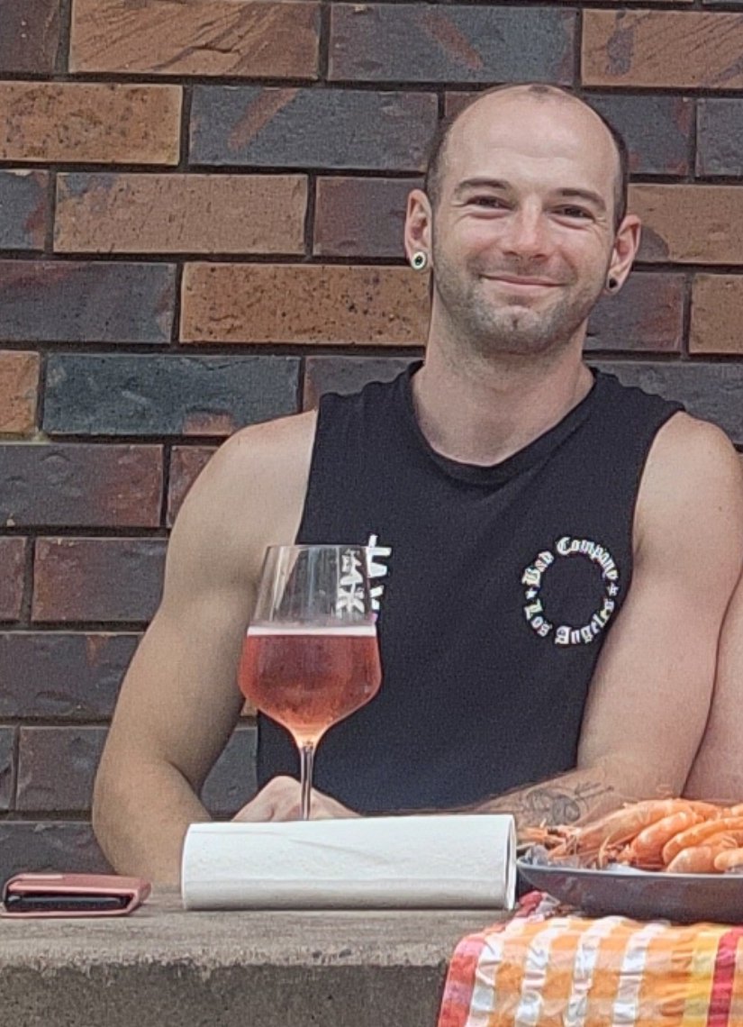 Josh from My Ultimate Hens smiling at an outdoor table with a glass of rosé and a plate of prawns