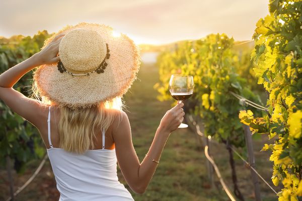 Gorgeous hen wearing white dress and hat while standing in a vineyard and holding a wine glass