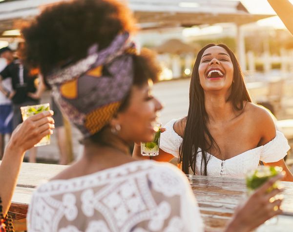 Two friends laughing with cocktails at a sunny beachside bar