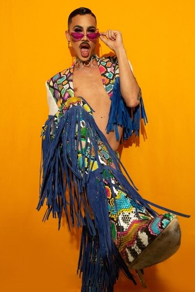 A drag queen in pink sunglasses and a fringed sequin outfit against an orange wall