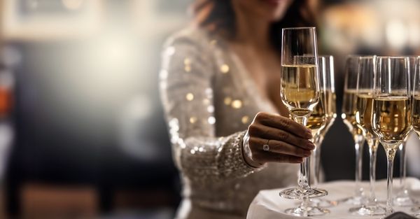 woman in sparkly dress picking up flute of bubbly from a table full of glasses of bubbly