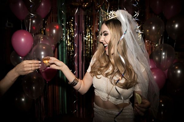 A bride to be in a veil and tiara laughing as she clinks shots among balloons