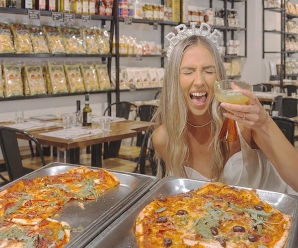 A bride in a tiara laughing with a cocktail over trays of fresh pizza