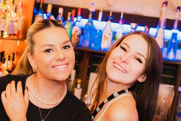 Two smiling women posing together in front of a lit cocktail bar shelf of bottles