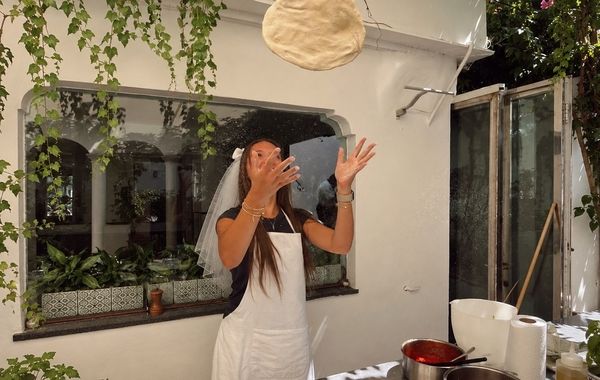 A bride in a veil and apron laughing as she tosses pizza dough in the air