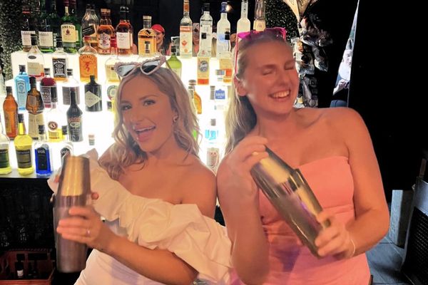 Two friends laughing as they hold up their cocktail shakers in front of a wall of bottles