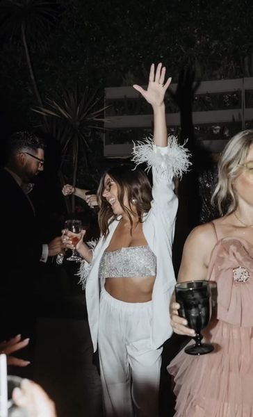 A bride to be in a white suit and feather cuffs cheering with friends at a night party