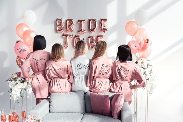 Hen and her best friends dressed in white and pink robes stand on their back and looking at the wall decorations in their hotel room