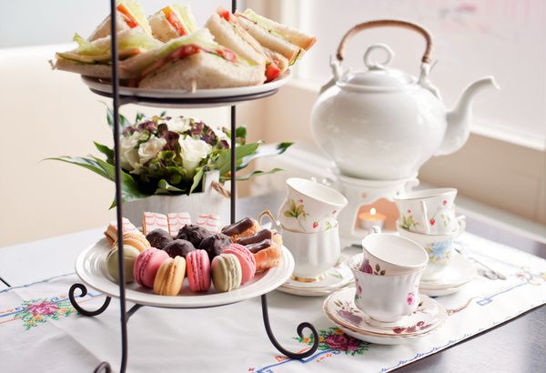 A high tea party with a tray of sweet pastries, macaroons, cupcakes, a cup of tea, teapots, and teacups set out on a white table