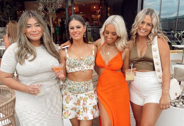 Four friends in summer dresses and florals smiling with cocktails on a sunny rooftop terrace
