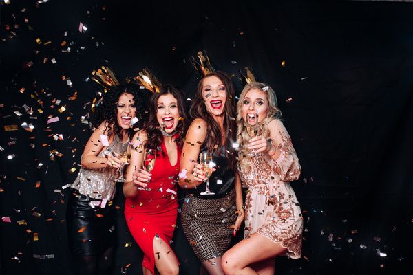 Four lovely ladies wearing fancy dresses and golden crowns smile in a group photo and have fun with confetti falling on them