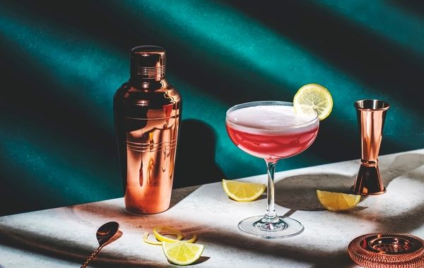 A cocktail drink garnished with lime slices on a white table with a shaker, citrus fruits, and jigger on the side