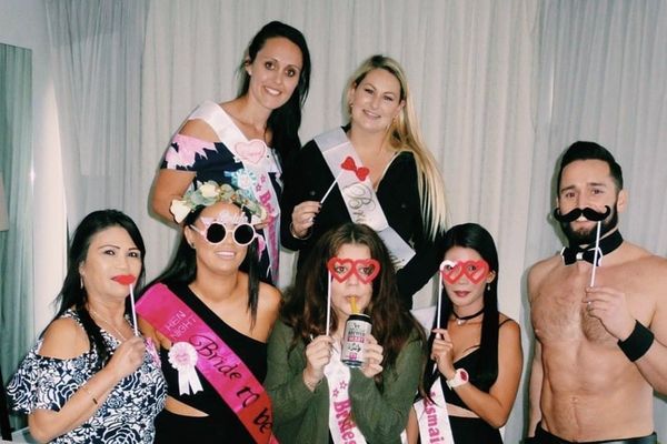 Hens in sashes hold photo props beside a shirtless waiter in a bow tie