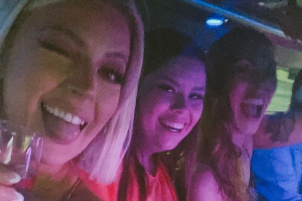 Three smiling women squeeze in for a selfie inside a limousine with drinks in hand