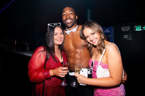 Topless waiter in a bow tie with two smiling hens holding glasses of bubbly