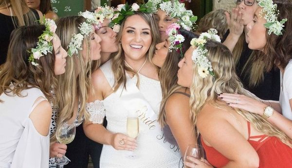 The bride in her sash laughing as her friends in flower crowns kiss her cheeks