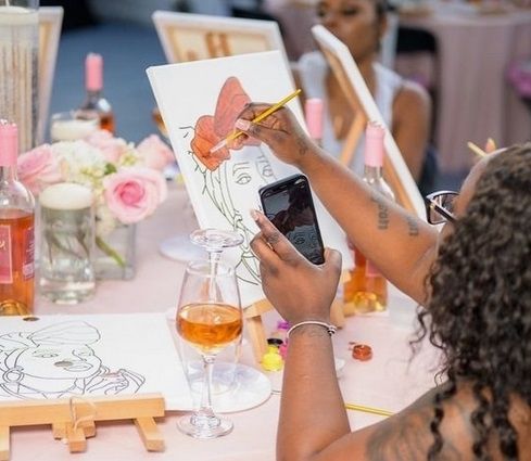 A hen photographing her canvas at a paint and sip table with wine and pink roses
