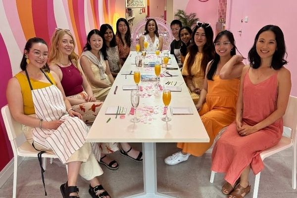 A hens group in aprons seated along a long studio table with glasses of bubbly