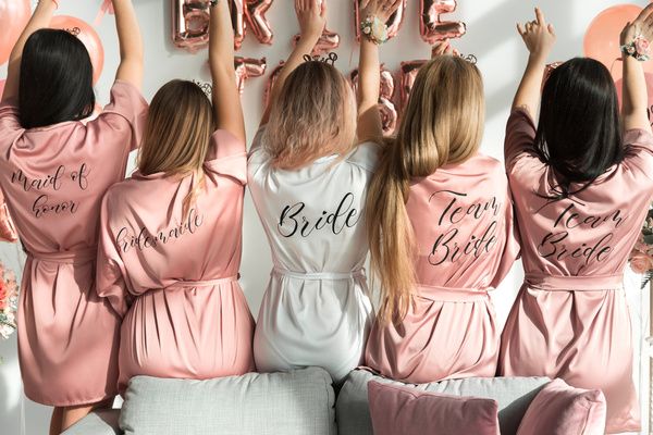 Five friends seen from behind in pink and white satin robes printed with Bride and Team Bride
