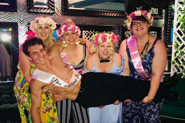 Hens in flower crowns and sashes laughing as they lift a topless waiter