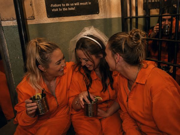 Three friends in orange prison jumpsuits laughing with drinks in tin cups beside a cell