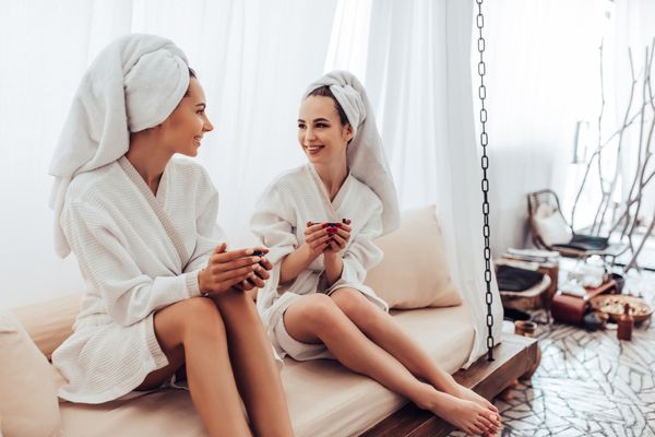 Two friends in white robes and towel turbans relaxing with drinks at a day spa