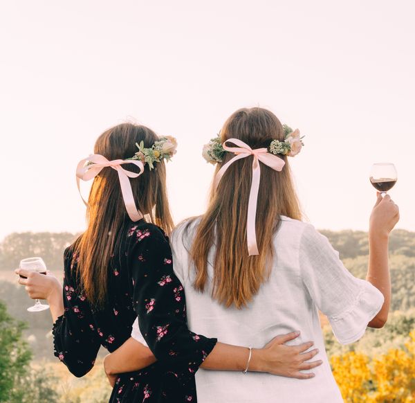 Two friends in flower crowns and pink ribbons with glasses of red wine, looking out over hills
