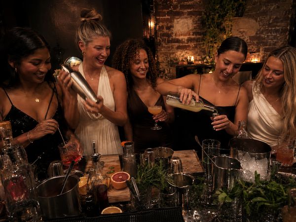 Five friends laughing as they shake and pour cocktails behind a bar