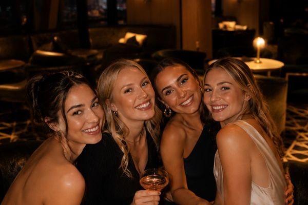 Four smiling women in evening dresses posing together in a warmly lit hotel bar