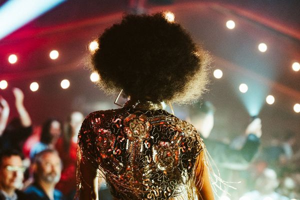 Cabaret performer in a sequinned fringe jacket walking past a seated audience