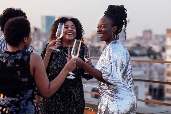 Three friends in sequins laughing and toasting on a rooftop