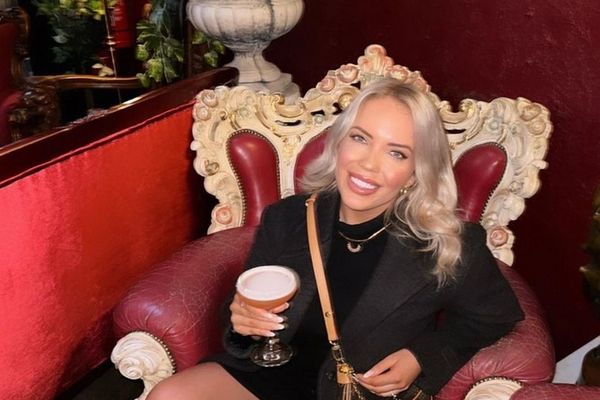 A smiling woman with a cocktail sits in an ornate red velvet chair