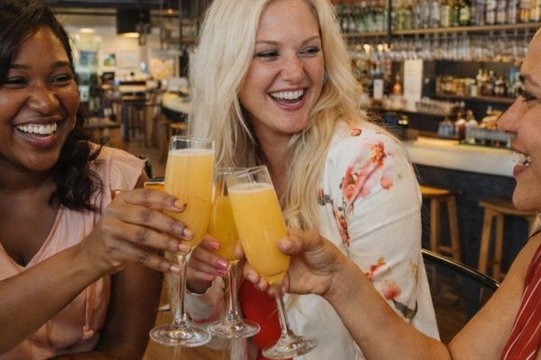 Three laughing women raising mimosas in a toast at a busy brunch bar