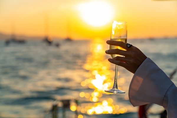 A hand holding a glass of bubbles up against a sunset over the water
