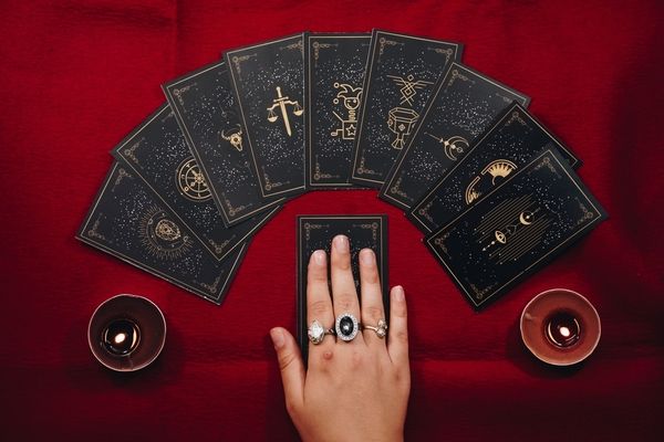 A hand with rings resting on a black tarot card, the deck fanned out on red velvet