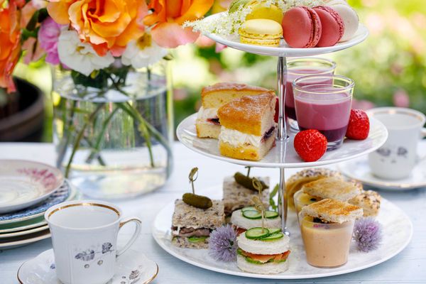 A tiered high tea stand with macarons, sandwiches and scones beside flowers and teacups