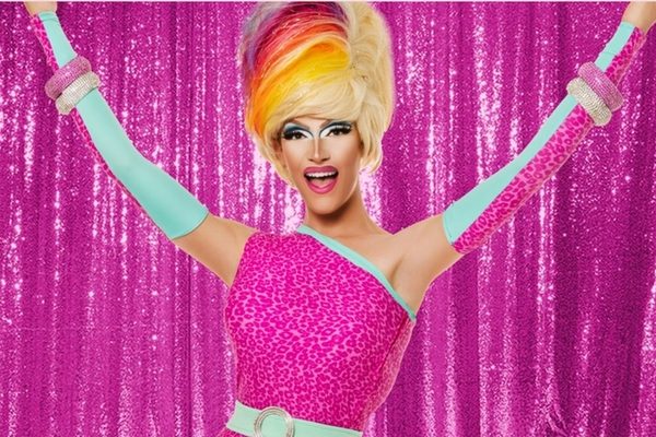A drag queen in a rainbow wig and pink sequins with her arms up