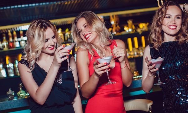 Three elegant ladies in red and black dresses stand at a bar counter and have fun during cocktail making class