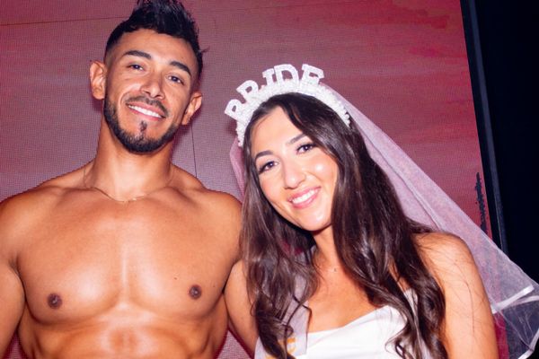 Bride in a veil and tiara poses with a shirtless waiter against a pink wall