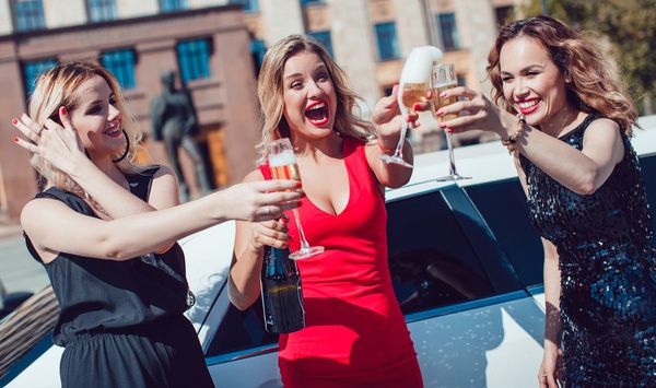 The hen is wearing a red dress and her best friends dressed in black dresses laughed and held champagne glasses in front of a white Hummer