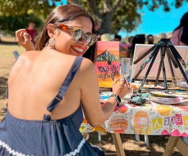 A friend in sunglasses smiling over her shoulder at an outdoor painting table in the sunshine