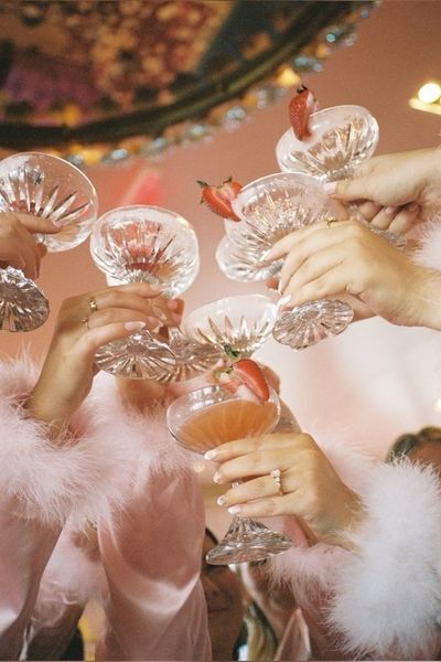 Hands in feather cuffs clinking coupes of pink cocktails