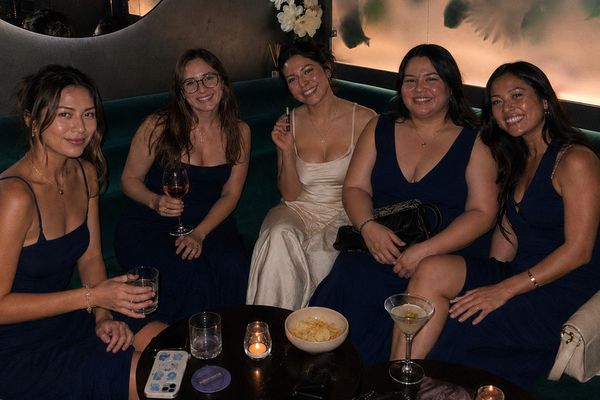 Five friends in dresses smiling on a green velvet banquette with cocktails and candles