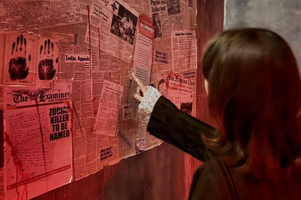 A woman pointing at a wall of old newspaper clues in a red-lit escape room