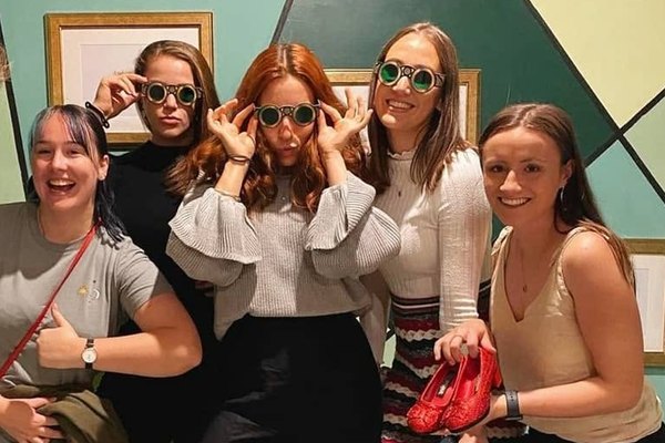 Five friends in an escape room pulling faces in round green glasses, one holding red sparkly shoes