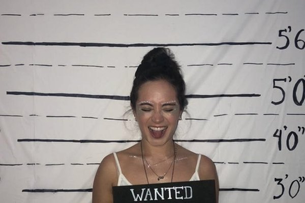 A laughing woman with face glitter holding a Wanted sign in front of a height chart
