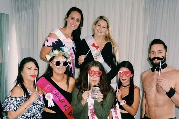 Hens in sashes holding photo props and posing with a shirtless waiter