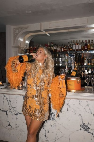A woman in an orange feathered dress drinking from a bottle of bubbles at a bar