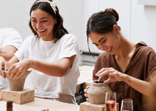 The bride in a white bow laughing with her friend as they shape their clay pieces, glasses of rose beside them